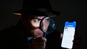 Homem com chapéu observando atentamente a tela de um celular com o aplicativo do Facebook aberto, segurando uma lupa em frente ao rosto em ambiente escuro, simbolizando investigação e curiosidade sobre perfis.
